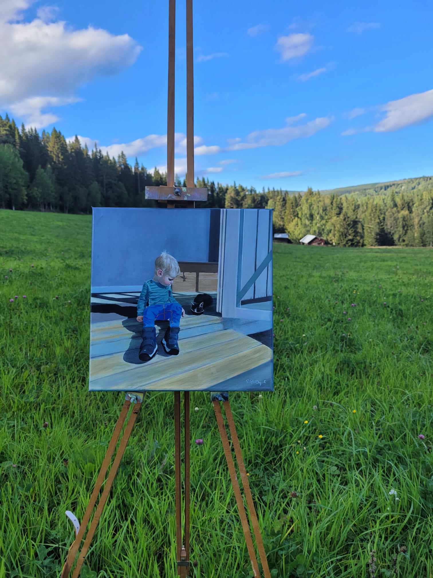 En liten pojke med napp i munnen tittar på en katt. Bilden är en vacker oljemålning. I bakgrunden syns skog och två hus. Oljemålningen står på ett staffli på en vall. Gräset är grönt och himlen blå.